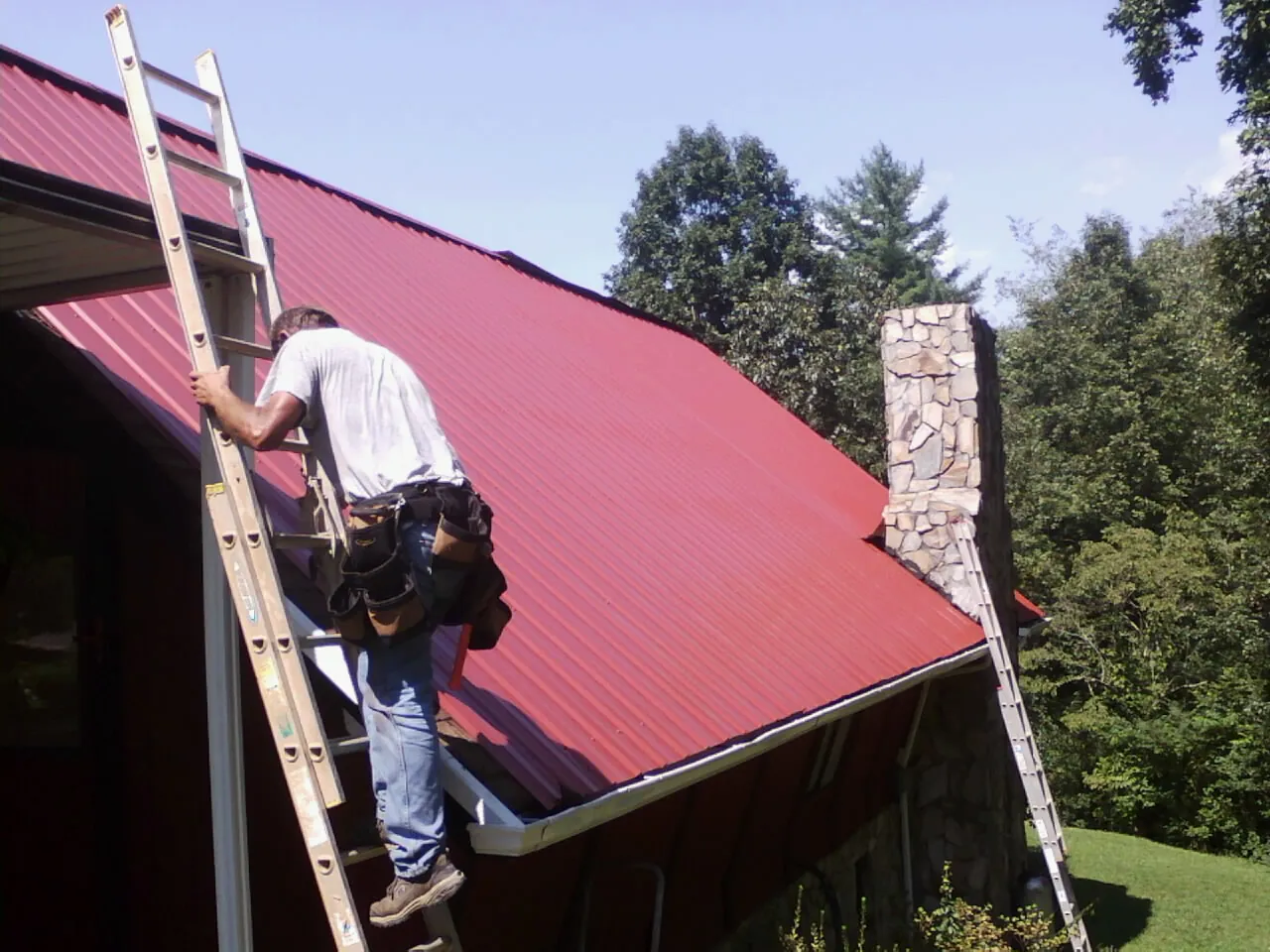 Roofer on a ladder beside a red metal roof and stone chimney — Soffit & Fascia Repair by Roofing Brothers in Malibu